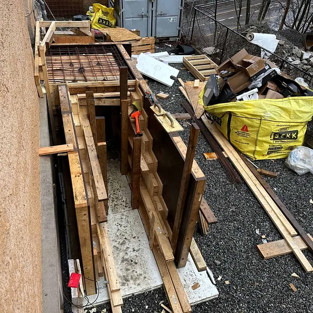 Forskaling og armering monteres for betongtrapp ved bygning, med materialer rundt.