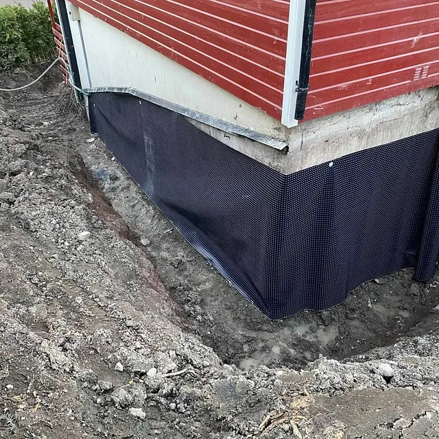 Grunnmur sikret med knotteplast under dreneringsarbeid langs bolig med utgravd grøft.