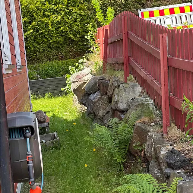 Eksisterende terreng med natursteinsmur og gjerde før oppstart av grave- og murarbeider.