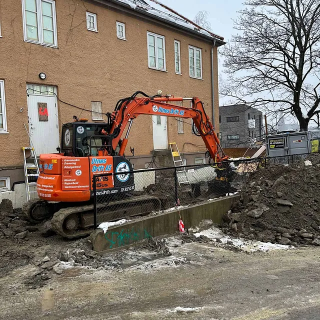Gravemaskin utfører grunnarbeid ved boligblokk, med steinmasser og snø langs tomten.
