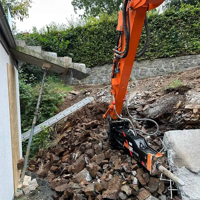 Gravemaskin med hydraulisk hammer utfører bergsprenging og oppgraving tett inntil bolig.