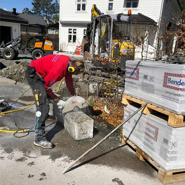 Arbeider skjærer granittkantstein med kappesag ved boligprosjekt, omgitt av maskiner og materialer.