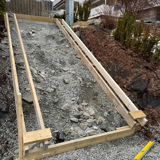 Forskalingsarbeid for ny betongtrapp pågår i skrånende terreng mellom bolig og gangvei.