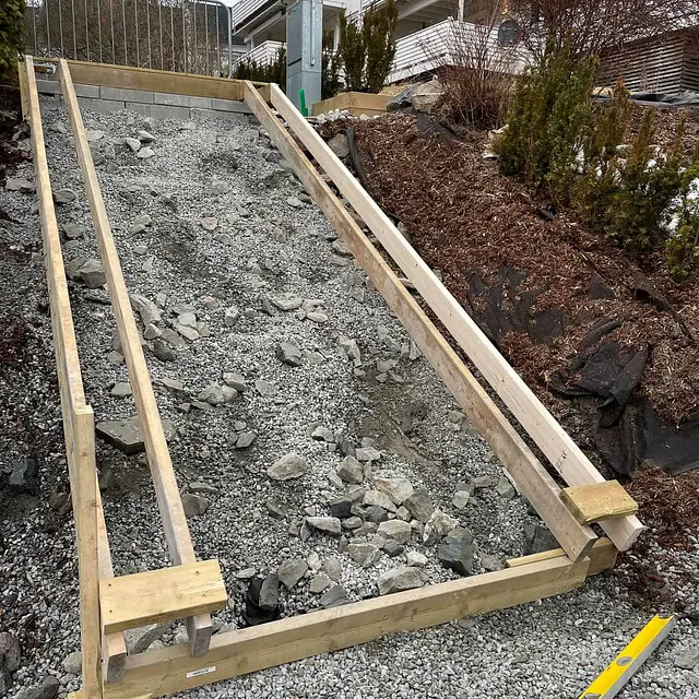 Forskaling til betongtrapp monteres i skråning mellom boliger, med grus og stein rundt.