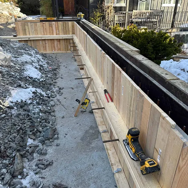Forskaling til betongmur bygges langs bolig, med verktøy og fundament klart for støping.