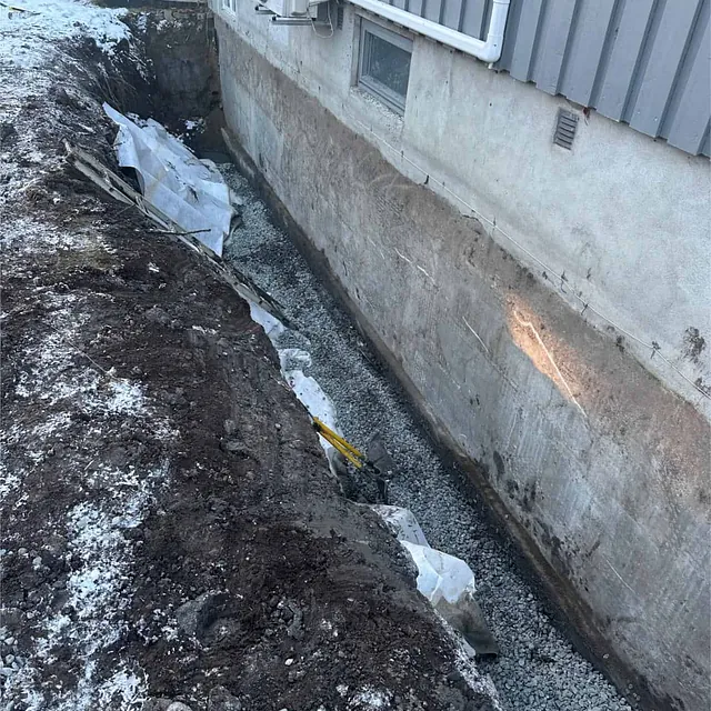 Utgravd grøft langs grunnmur klargjort med pukk som grunnlag for nytt dreneringssystem.