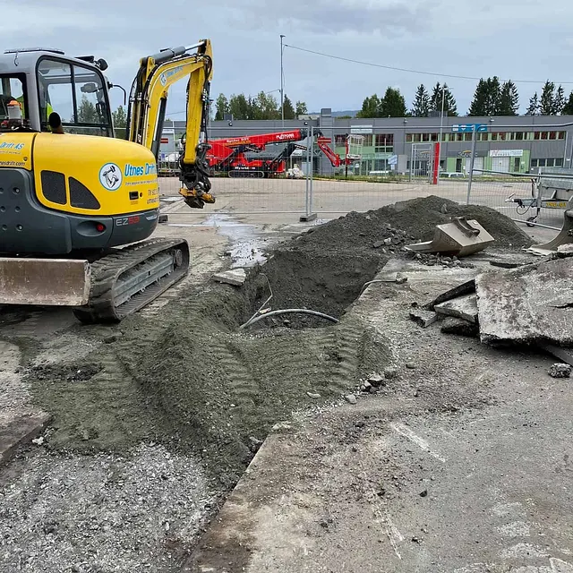 Gravemaskin graver grøft for infrastrukturarbeid på næringstomt med bygg og utstyr rundt.