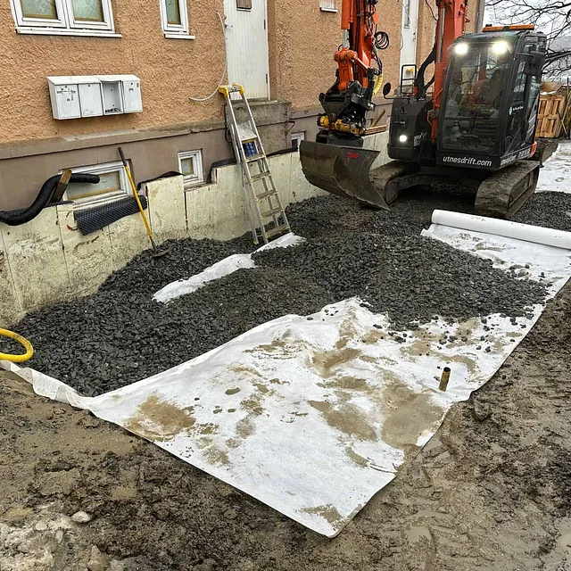 Dreneringsarbeid langs grunnmur med duk, pukk og gravemaskin klargjort for tilbakefylling.