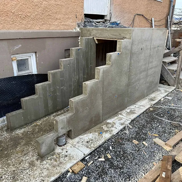 Støpt betongtrapp under oppføring ved husvegg, med drenering og byggearbeid rundt.