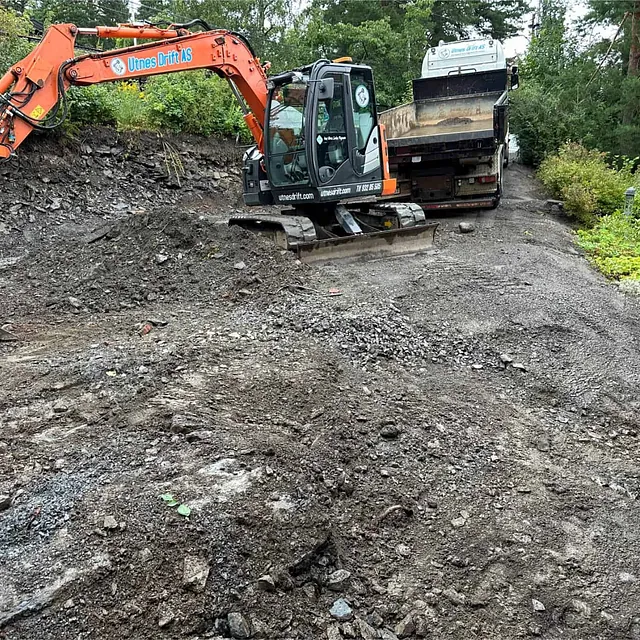 Gravemaskin og lastebil utfører masseflytting og terrengarbeid på byggeplass ved bolig.