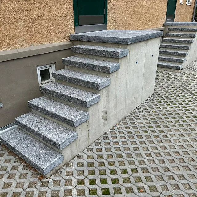 Ferdigstilt betongtrapp med granitttrinn ved inngangsparti, omgitt av belegningsstein.