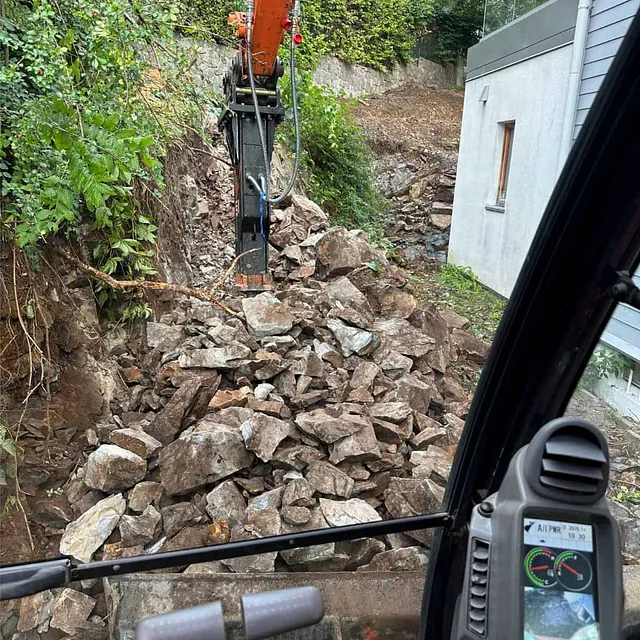 Gravemaskin med hydraulisk hammer utfører bergarbeid langs boligens sideområde.