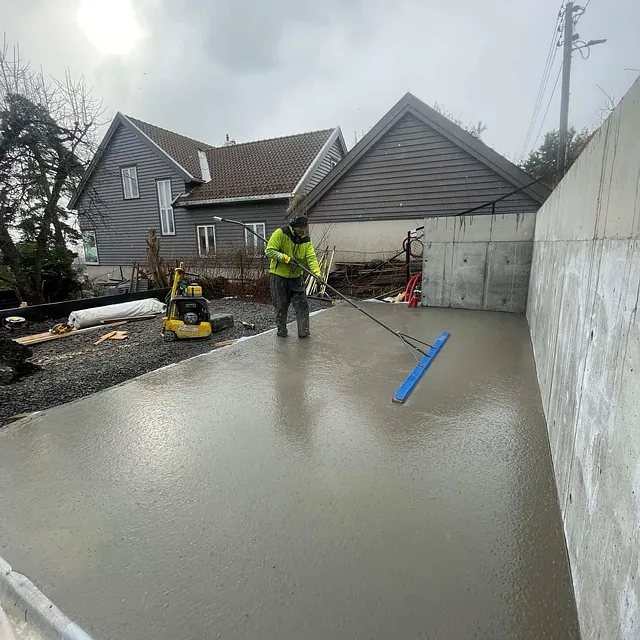Arbeider glatter fersk betongplate ved støttemur, som del av fundament- og utearbeid.