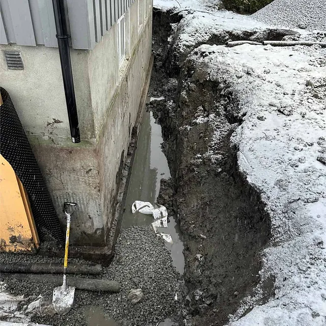 Utgraving langs grunnmur med pukk og vannansamling før ferdigstillelse av dreneringen.