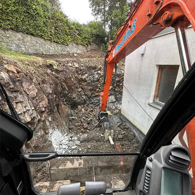 Gravemaskin med hydraulisk hammer utfører bergsprenging og oppgraving tett inntil bolig.