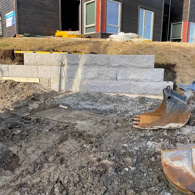Natursteinsmur under oppføring med flere steinlag montert for terrengstøtte ved bolig.