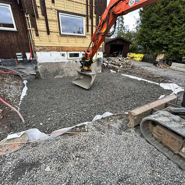 Pukk fordeles og avrettes med gravemaskin som bærelag for nytt fundament ved boligen.