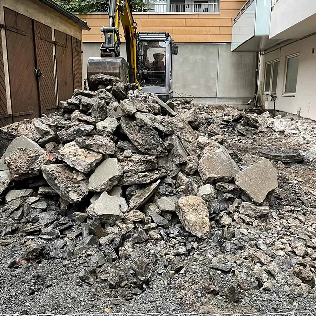 Oppbrutt asfalt og betong fjernes med gravemaskin under rehabilitering av gårdsplass.