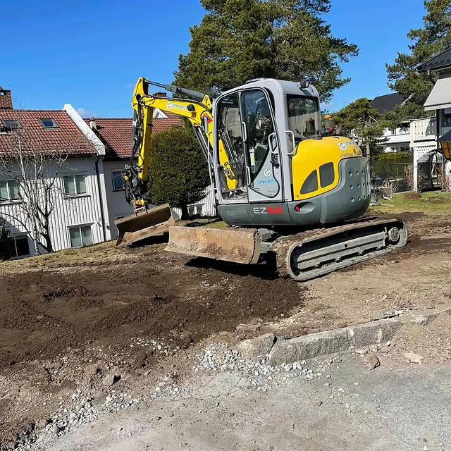 Gravemaskin utfører terrengarbeid på tomt med boliger, trær og klargjøring for hageanlegg.